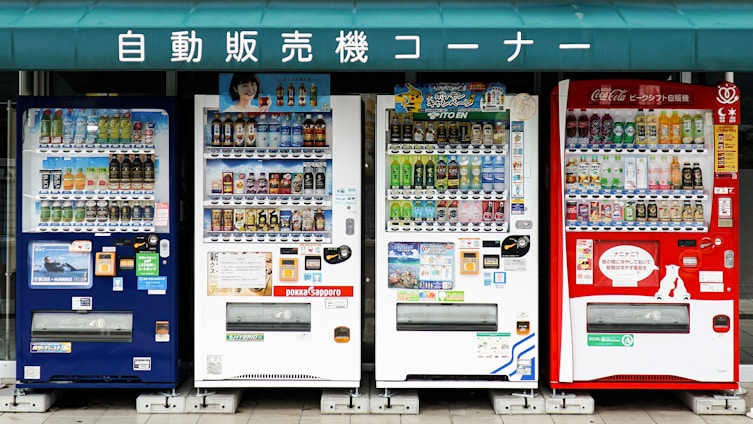 A row of four vending machines is displayed, each offering a variety of beverages. The machines are colorful, with numerous bottles and cans visible through the glass. Above the machines, there is a green sign with white Japanese text.