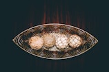 A decorative metal tray with an intricate swirled design holds four spherical objects that resemble natural woven orbs. The objects are arranged in a linear fashion, each with unique textures and colors. The background consists of a rich, dark wood surface, enhancing the rustic and artistic feel of the composition.