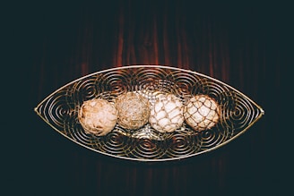 A decorative metal tray with an intricate swirled design holds four spherical objects that resemble natural woven orbs. The objects are arranged in a linear fashion, each with unique textures and colors. The background consists of a rich, dark wood surface, enhancing the rustic and artistic feel of the composition.