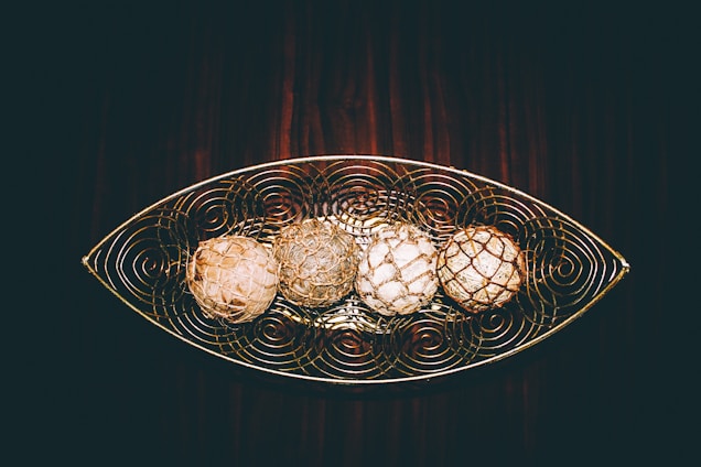 A decorative metal tray with an intricate swirled design holds four spherical objects that resemble natural woven orbs. The objects are arranged in a linear fashion, each with unique textures and colors. The background consists of a rich, dark wood surface, enhancing the rustic and artistic feel of the composition.