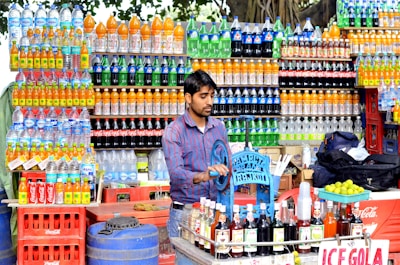 A street vendor is preparing drinks at a colorful stall filled with various bottles of soda and water. The shelves are lined with bottles of vibrant colors, including orange, green, and blue. The vendor is using a manual machine to prepare beverages, and a sign for 'Ice Gola' is visible. Limes and syrup bottles are on the counter.