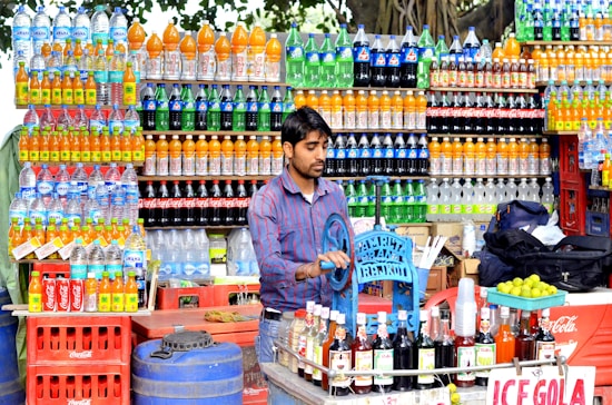 A street vendor is preparing drinks at a colorful stall filled with various bottles of soda and water. The shelves are lined with bottles of vibrant colors, including orange, green, and blue. The vendor is using a manual machine to prepare beverages, and a sign for 'Ice Gola' is visible. Limes and syrup bottles are on the counter.