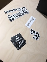 A collection of Unsplash stickers are arranged on a light wooden surface. The stickers include the Unsplash logo in various forms: two black square stickers with the logo, several white rectangular ones, and smaller square stickers with only the logo. The stickers are casually placed, creating an informal and creative mood.