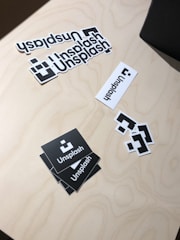 A collection of Unsplash stickers are arranged on a light wooden surface. The stickers include the Unsplash logo in various forms: two black square stickers with the logo, several white rectangular ones, and smaller square stickers with only the logo. The stickers are casually placed, creating an informal and creative mood.