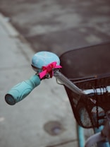 A close-up of a bicycle handlebar with a light blue grip and a bell. The bell is decorated with a vibrant pink ribbon. Attached to the handlebar is a black mesh basket. The background is out of focus, showing a concrete pavement.