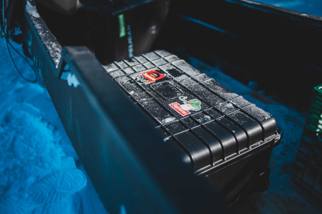 A sturdy black plastic box with ridged edges and a couple of colorful stickers on top is placed in the back of an open vehicle or trailer. The surface is lightly dusted with snow, and the surrounding area also has snow, with tracks visible. The lighting gives the scene a cool, wintery ambiance.