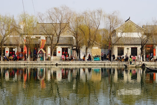 A tranquil riverside scene features traditional buildings with tiled roofs and bare willow trees. People stroll along the walkway, some sitting on benches near the water. Decorative red banners are visible on the buildings, adding a festive touch. The calm water reflects the buildings and trees, creating a serene atmosphere.