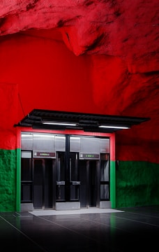 A modern setting with two elevator doors positioned against a wall. The upper portion of the wall features a vibrant red color with a rocky texture, while the lower portion is painted in green. Smooth reflective flooring adds a sleek look to the space.