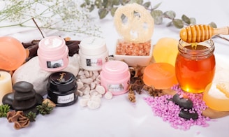 A variety of spa and skincare items are arranged aesthetically, including jars of cream, bars of soap, a glass jar filled with honey, and pink salt displayed in a small bowl. Dried herbs and smooth black stones enhance the natural and calming theme.