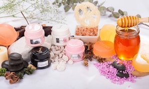 A variety of spa and skincare items are arranged aesthetically, including jars of cream, bars of soap, a glass jar filled with honey, and pink salt displayed in a small bowl. Dried herbs and smooth black stones enhance the natural and calming theme.