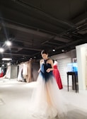 A person wearing a gradient ombre gown, ranging from dark blue at the top to a light beige at the bottom, stands in a well-lit showroom filled with a variety of elegant dresses in various colors hanging on racks. The setting appears upscale and sophisticated, with a modern interior design featuring spotlights on the ceiling.