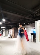 A person wearing a gradient ombre gown, ranging from dark blue at the top to a light beige at the bottom, stands in a well-lit showroom filled with a variety of elegant dresses in various colors hanging on racks. The setting appears upscale and sophisticated, with a modern interior design featuring spotlights on the ceiling.