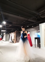 A person wearing a gradient ombre gown, ranging from dark blue at the top to a light beige at the bottom, stands in a well-lit showroom filled with a variety of elegant dresses in various colors hanging on racks. The setting appears upscale and sophisticated, with a modern interior design featuring spotlights on the ceiling.