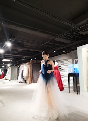 A person wearing a gradient ombre gown, ranging from dark blue at the top to a light beige at the bottom, stands in a well-lit showroom filled with a variety of elegant dresses in various colors hanging on racks. The setting appears upscale and sophisticated, with a modern interior design featuring spotlights on the ceiling.