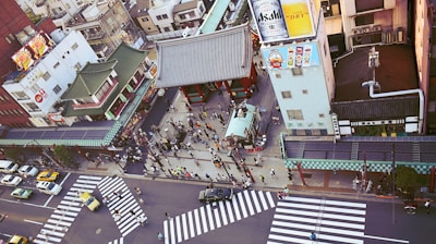 An aerial view of a busy urban area showcasing traditional Japanese architecture with temple buildings and a street filled with people and vehicles. There's a large building displaying advertisements, including a beer ad and animated characters.