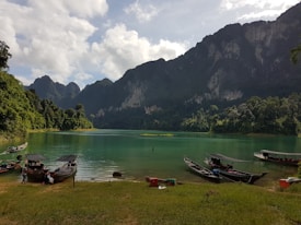 A serene lake surrounded by lush green forests and towering mountain ranges under a partly cloudy sky. Longtail boats are moored along the shore, with a few people nearby. The water is a clear turquoise, reflecting the vibrant greenery and rocky cliffs.