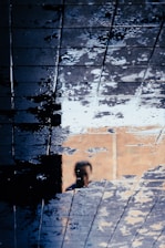 A reflection of a person is seen on a wet tiled surface, creating an abstract and distorted effect. The tiles are covered with water, causing a mixture of light and dark patches across the surface.