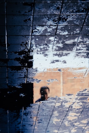 A reflection of a person is seen on a wet tiled surface, creating an abstract and distorted effect. The tiles are covered with water, causing a mixture of light and dark patches across the surface.