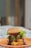 A juicy hamburger stacked with layers of cheese, fresh lettuce, tomato slices, and drizzled with barbecue sauce sits on an orange plate. The sesame seed bun tops the decadent layers, suggesting a hearty and satisfying meal. The blurred background puts the focus entirely on the appetizing burger.