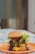 A juicy hamburger stacked with layers of cheese, fresh lettuce, tomato slices, and drizzled with barbecue sauce sits on an orange plate. The sesame seed bun tops the decadent layers, suggesting a hearty and satisfying meal. The blurred background puts the focus entirely on the appetizing burger.