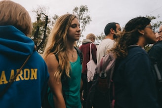 A group of people, mostly young, are walking outdoors. Some individuals are wearing backpacks, and the trees in the background suggest a park or a natural setting. The overall atmosphere is casual as people appear to be socializing.