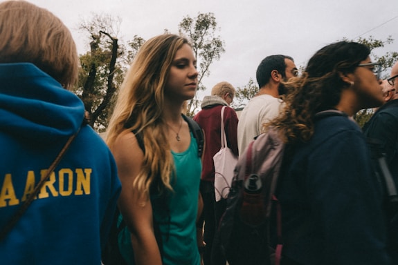 A group of people, mostly young, are walking outdoors. Some individuals are wearing backpacks, and the trees in the background suggest a park or a natural setting. The overall atmosphere is casual as people appear to be socializing.