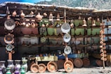 A stall displaying a variety of ceramic pottery items, including plates, vases, and traditional tagine pots. The pottery is arranged neatly on wooden shelves with several items hanging above. The background is a makeshift structure using cloth or tarp, and the scene has a rustic, artisanal feel.