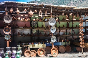 A stall displaying a variety of ceramic pottery items, including plates, vases, and traditional tagine pots. The pottery is arranged neatly on wooden shelves with several items hanging above. The background is a makeshift structure using cloth or tarp, and the scene has a rustic, artisanal feel.