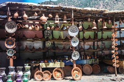A stall displaying a variety of ceramic pottery items, including plates, vases, and traditional tagine pots. The pottery is arranged neatly on wooden shelves with several items hanging above. The background is a makeshift structure using cloth or tarp, and the scene has a rustic, artisanal feel.