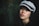 A young woman gazes softly into the camera, wearing a textured gray cap with a black band. Her gentle expression is highlighted by soft lighting that accentuates her features against a dark, leafy background.