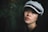 A young woman gazes softly into the camera, wearing a textured gray cap with a black band. Her gentle expression is highlighted by soft lighting that accentuates her features against a dark, leafy background.