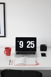 A minimalist workspace with a white desk features a laptop displaying a large digital clock reading 9:25 AM. To the left of the laptop is a red coffee mug, and in front of it lies an open notebook with a pen and pencil beside it. On the right side, a small black speaker or device and a smartphone are placed. The background shows a framed piece of art on the wall.