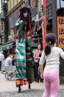 A person dressed in an elaborate costume with striped sleeves, fur accents, and long dreadlocks, stands on stilts on a bustling street. The costume features bright red, green, and gray colors with horns on the head. A person dressed casually in a plaid shirt is standing nearby, smiling, while a child in polka dot leggings looks on.