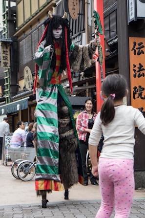 A person dressed in an elaborate costume with striped sleeves, fur accents, and long dreadlocks, stands on stilts on a bustling street. The costume features bright red, green, and gray colors with horns on the head. A person dressed casually in a plaid shirt is standing nearby, smiling, while a child in polka dot leggings looks on.
