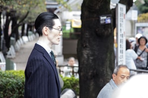 A person in profile wearing a dark pinstripe suit stands outdoors in a park-like setting with trees and blurred people in the background. The individual is facing slightly to the left, wearing glasses, with neatly combed dark hair. The background includes greenery and a mix of people sitting or standing around.