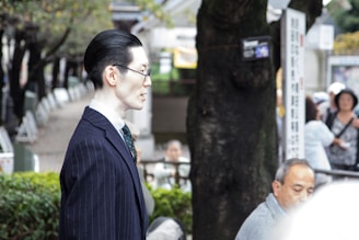 A person in profile wearing a dark pinstripe suit stands outdoors in a park-like setting with trees and blurred people in the background. The individual is facing slightly to the left, wearing glasses, with neatly combed dark hair. The background includes greenery and a mix of people sitting or standing around.