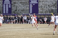 A group of female lacrosse players are actively participating in a game. One player is in the foreground about to catch or throw a ball with her stick, while others are either playing or watching intently. Spectators and coaches stand on the sidelines next to a bench area. Banners on the wall behind them display past championships and achievements.