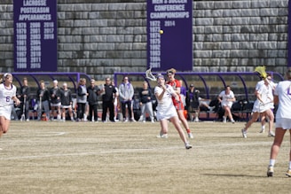 A group of female lacrosse players are actively participating in a game. One player is in the foreground about to catch or throw a ball with her stick, while others are either playing or watching intently. Spectators and coaches stand on the sidelines next to a bench area. Banners on the wall behind them display past championships and achievements.