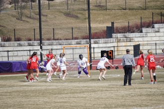 A group of female athletes is playing lacrosse on a grassy field. Players are wearing helmets and uniforms, with one team in red and the other in white. A referee dressed in black and white stripes watches the action. The scene includes a lacrosse goal with a net and a scoreboard showing the number nine. The backdrop includes a stone wall and sparse vegetation.