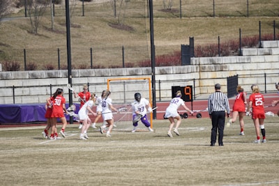 A group of female athletes is playing lacrosse on a grassy field. Players are wearing helmets and uniforms, with one team in red and the other in white. A referee dressed in black and white stripes watches the action. The scene includes a lacrosse goal with a net and a scoreboard showing the number nine. The backdrop includes a stone wall and sparse vegetation.