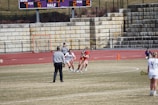 A lacrosse game in progress on a grassy field with players wearing uniforms. A referee in a striped shirt is overseeing the game. The scene includes a scoreboard in the background displaying the current score and time, and stone bleachers filled with spectators.