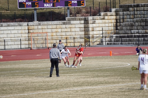 A lacrosse game in progress on a grassy field with players wearing uniforms. A referee in a striped shirt is overseeing the game. The scene includes a scoreboard in the background displaying the current score and time, and stone bleachers filled with spectators.