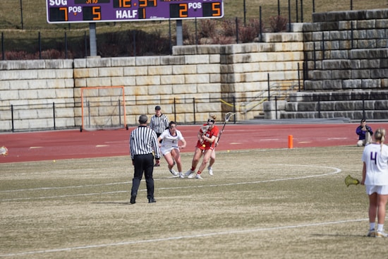 A lacrosse game in progress on a grassy field with players wearing uniforms. A referee in a striped shirt is overseeing the game. The scene includes a scoreboard in the background displaying the current score and time, and stone bleachers filled with spectators.