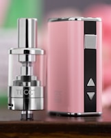 A close-up view of a sleek, metallic vaping device with a glossy finish and a pink and black color scheme. The device features buttons and a clear tank, indicating its function as an electronic cigarette.