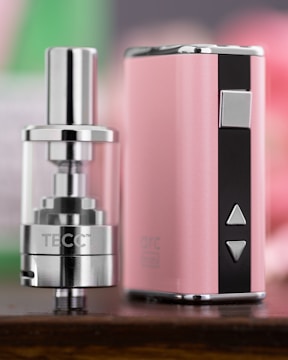 A close-up view of a sleek, metallic vaping device with a glossy finish and a pink and black color scheme. The device features buttons and a clear tank, indicating its function as an electronic cigarette.