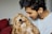 A young man is affectionately holding a fluffy, curly-haired dog. The man has a gentle smile on his face, indicating a close bond with the pet. The setting appears to be indoors, with a red couch partially visible in the background.