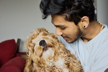 A young man is affectionately holding a fluffy, curly-haired dog. The man has a gentle smile on his face, indicating a close bond with the pet. The setting appears to be indoors, with a red couch partially visible in the background.
