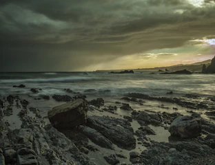 A dramatic sky filled with dark storm clouds and streaks of sunlight breaking through over a rugged coastline.