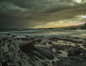 A dramatic sky filled with dark storm clouds and streaks of sunlight breaking through over a rugged coastline.