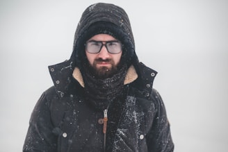 A person wearing a winter coat and a hood stands in a snowy environment. The individual's face is partially covered by a scarf, and they are wearing glasses. Snowflakes are visible on the clothing and in the air, indicating cold weather.